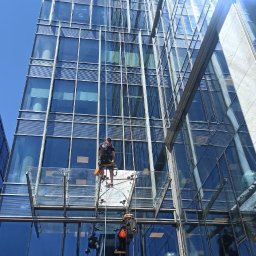 SkyCloud Ruslan Okilka - Dwóch alpinistów na platformie myje szklaną fasadę wieżowca w słoneczny dzień. Widok z dołu, podkreślający wysokość i nowoczesną architekturę budynku.