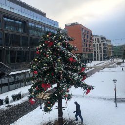 SkyCloud Ruslan Okilka - Ośnieżone otoczenie biurowca z dużą, udekorowaną choinką. Mężczyzna w ciemnej kurtce i niebieskich spodniach przechadza się obok drzewka. Zimowy krajobraz miejski.