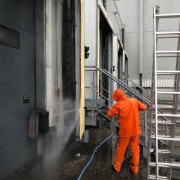 SkyCloud Ruslan Okilka - Pracownik w pomarańczowym kombinezonie czyści elewację hali przemysłowej myjką ciśnieniową. Widoczna drabina i fragmenty elewacji z rampami załadunkowymi.