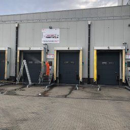 SkyCloud Ruslan Okilka - Mycie bram przemysłowych z użyciem myjki ciśnieniowej przez pracownika w kombinezonie ochronnym. Widoczne trzy bramy, drabina i woda pod ciśnieniem.