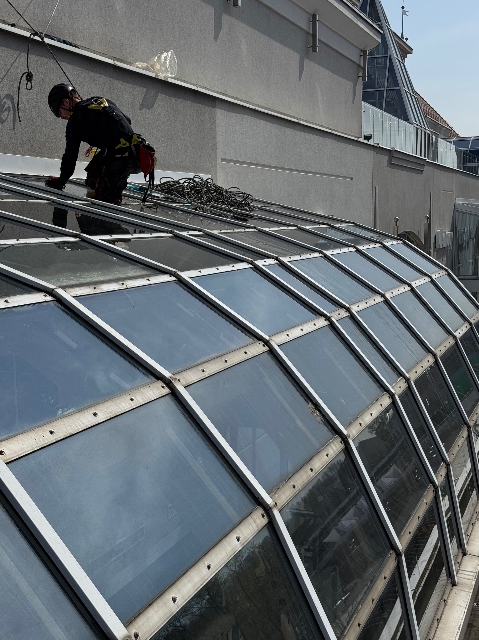 Pracownik w uprzęży, na szklanym dachu, podczas czyszczenia. Widoczne liny, kask i narzędzia. Ujęcie z dołu, podkreślające wysokość i specyfikę pracy.