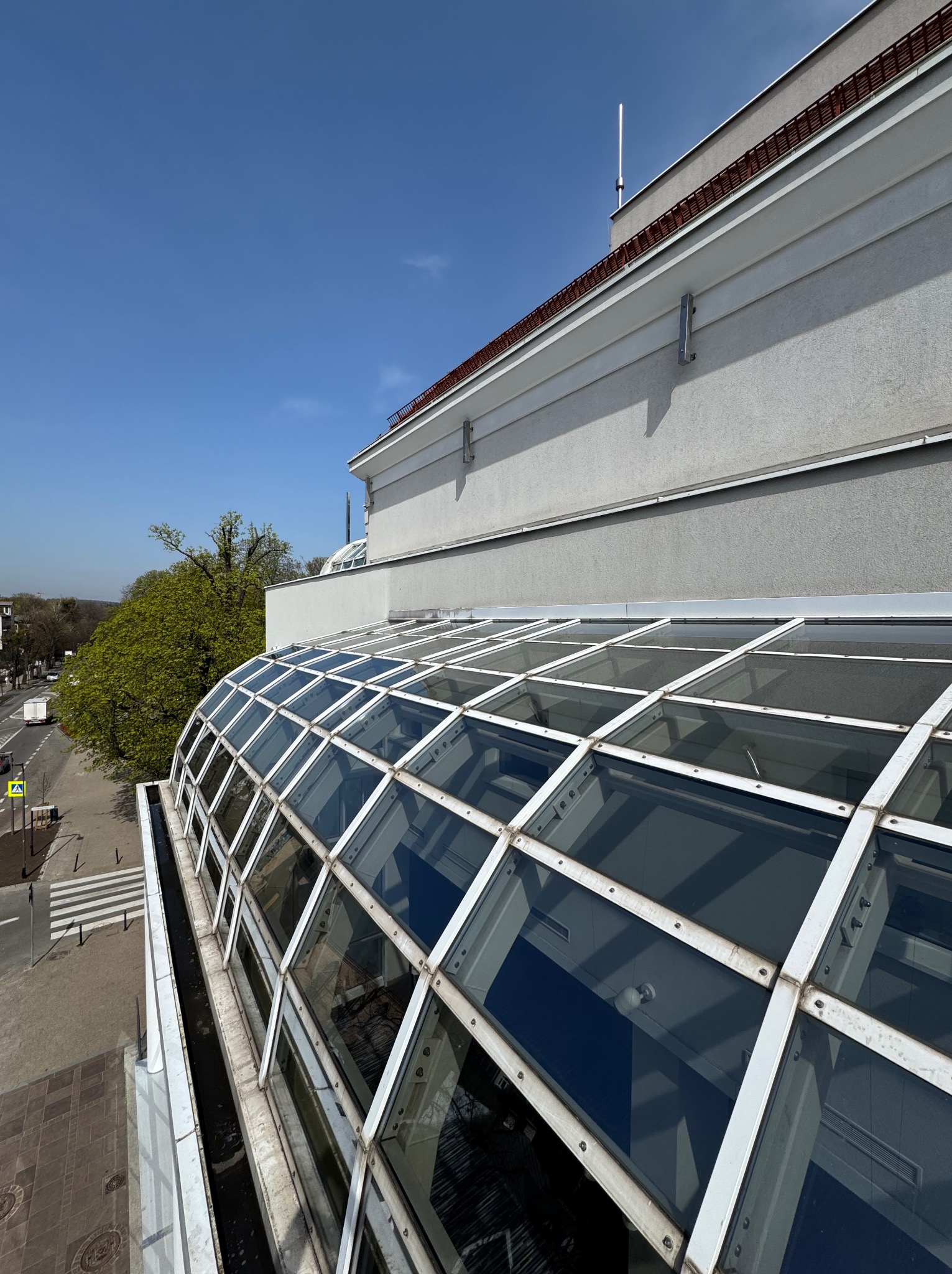 Szklany dach budynku z widocznymi zabrudzeniami i mchem, ujęcie z góry, fragment elewacji i rynny, błękitne niebo w tle. Widok na ulicę i zieleń.