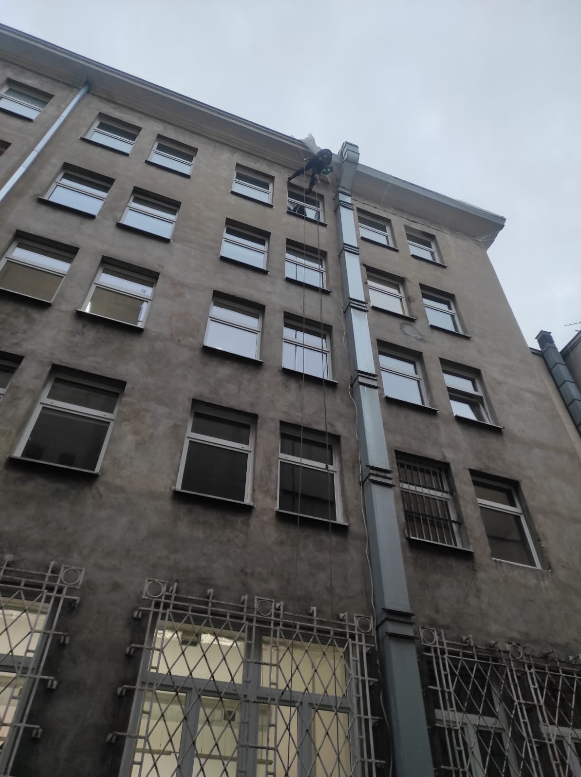 Pracownik na linie zabezpieczonej, wykonujący prace na wysokości przy budynku z szarą elewacją i licznymi oknami. Widok z dołu, pochmurne niebo.