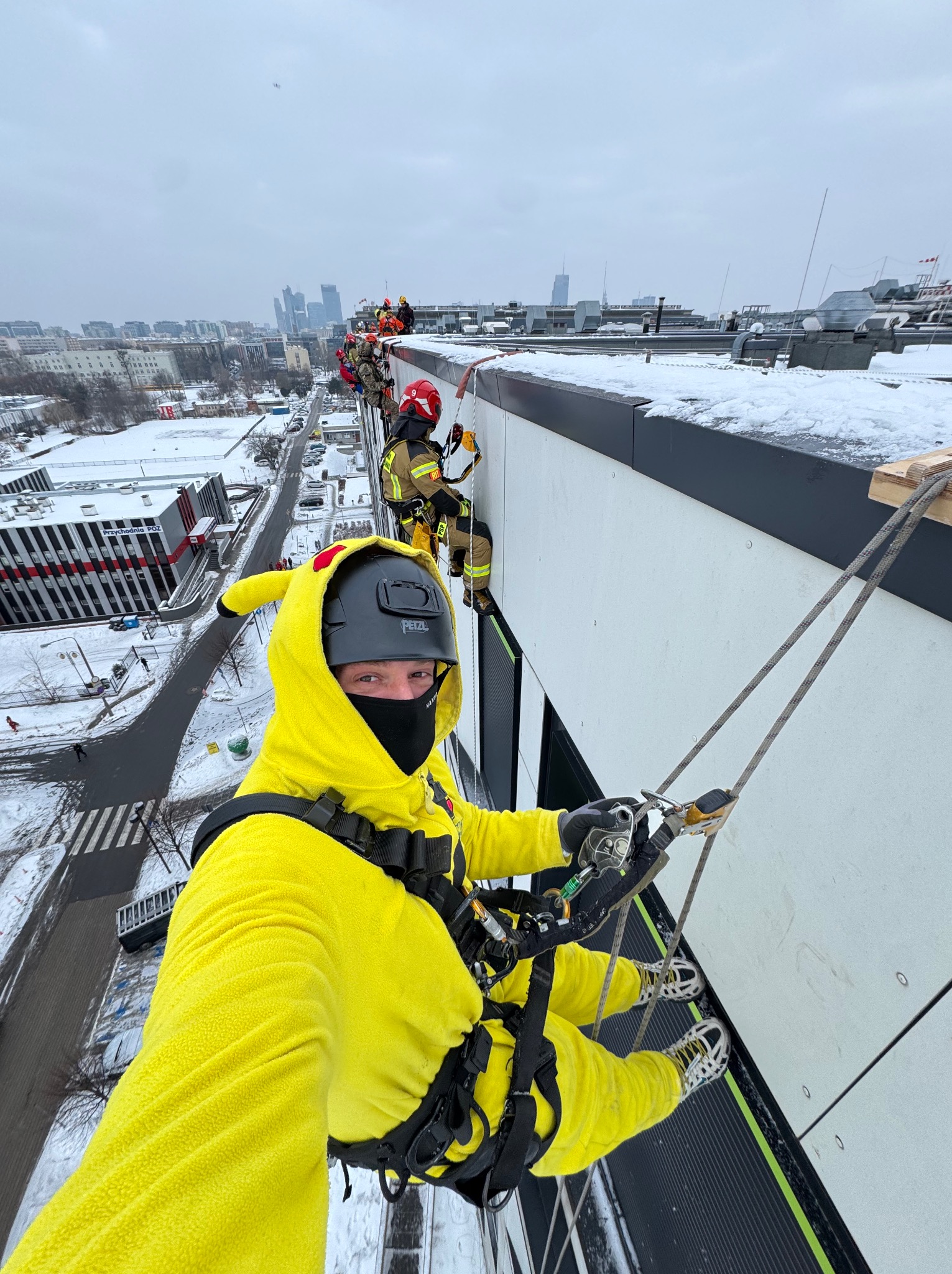 Osoba w kostiumie Pikachu, w kasku i masce, na linie podczas prac wysokościowych na budynku w zimowej scenerii miejskiej. W tle inni pracownicy i panorama miasta.