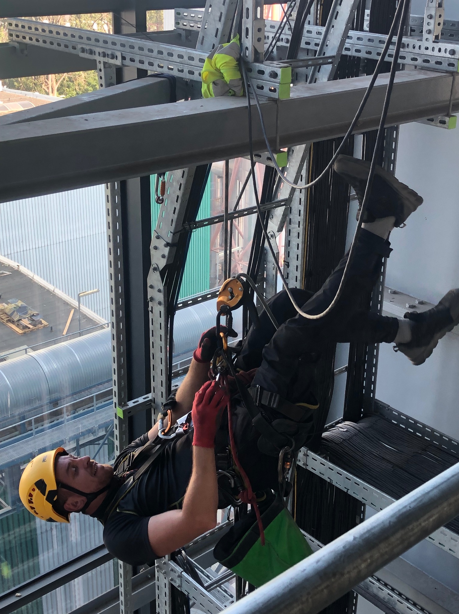 Pracownik w kasku i uprzęży, zawieszony na linach, wykonuje prace na wysokości na tle konstrukcji stalowej i budynku. Widoczny drugi pracownik w oddali.