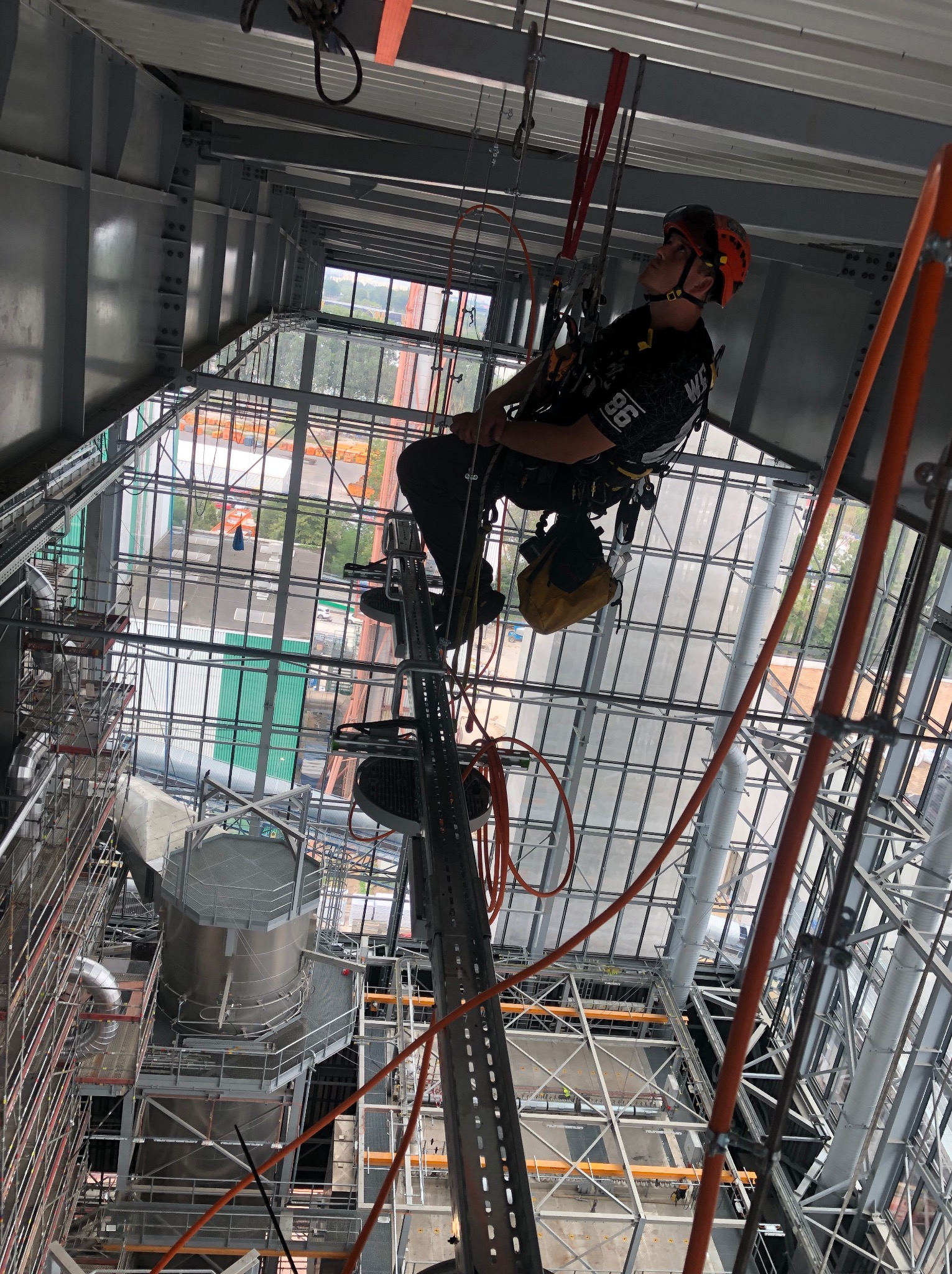 Pracownik w kasku, zabezpieczony linami, na wysokości wewnątrz konstrukcji stalowej z przeszklonym dachem, wykonuje prace na wysokościach.