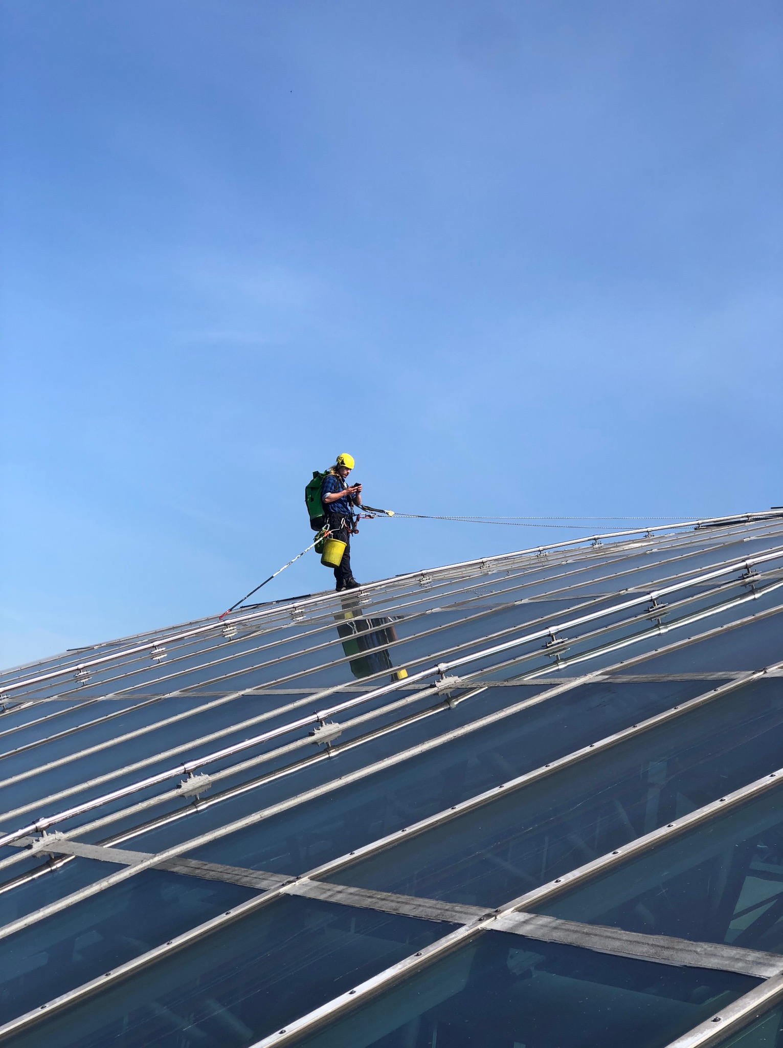 Pracownik w kasku i uprzęży, z zielonym plecakiem, czyści szklany dach z użyciem specjalistycznego sprzętu. Widok z dołu na błękitne niebo.