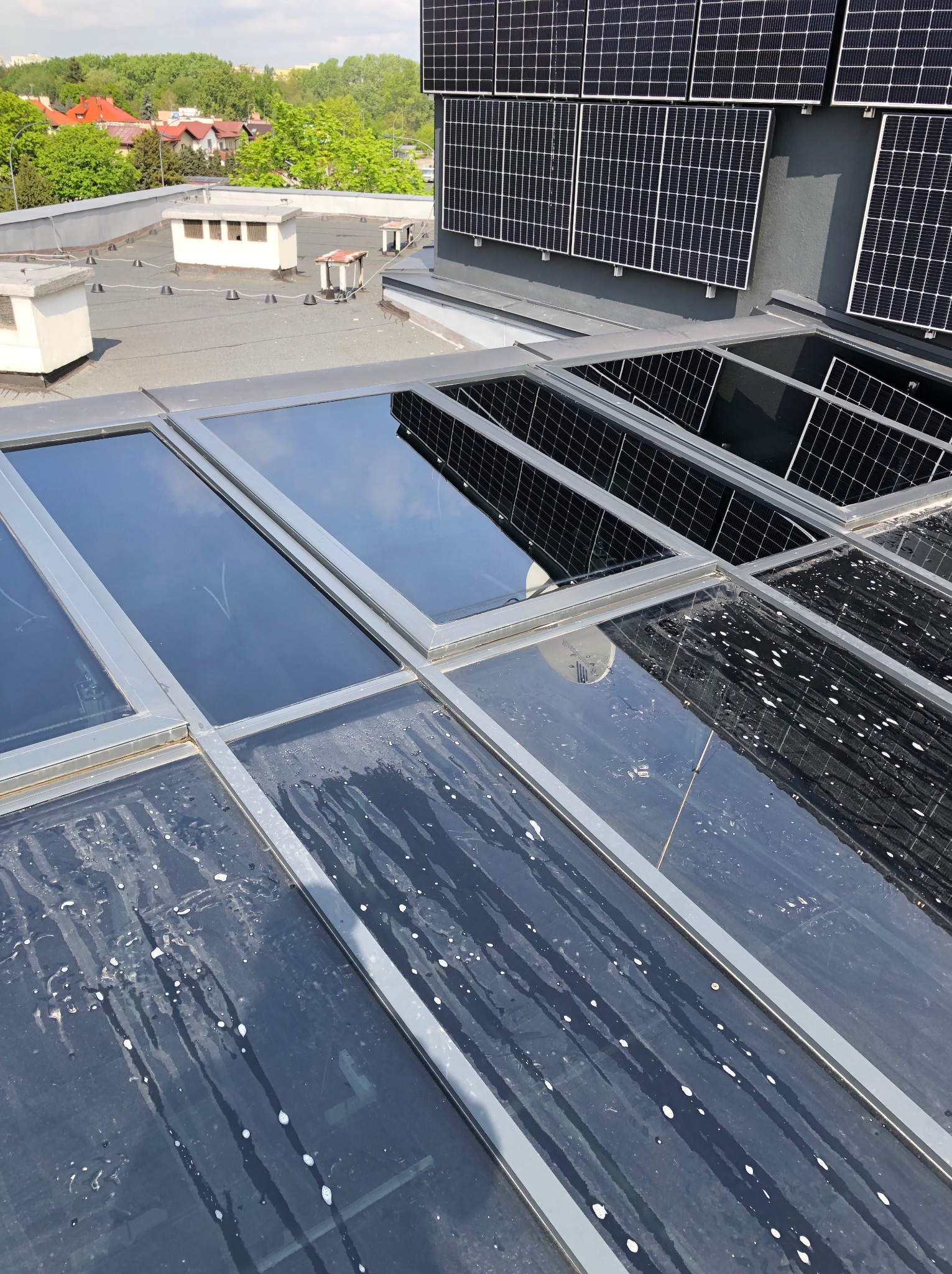 Zabrudzone okna dachowe na płaskim dachu z panelami słonecznymi w tle. Widoczne smugi i plamy po deszczu. Ujęcie z góry, w słoneczny dzień.