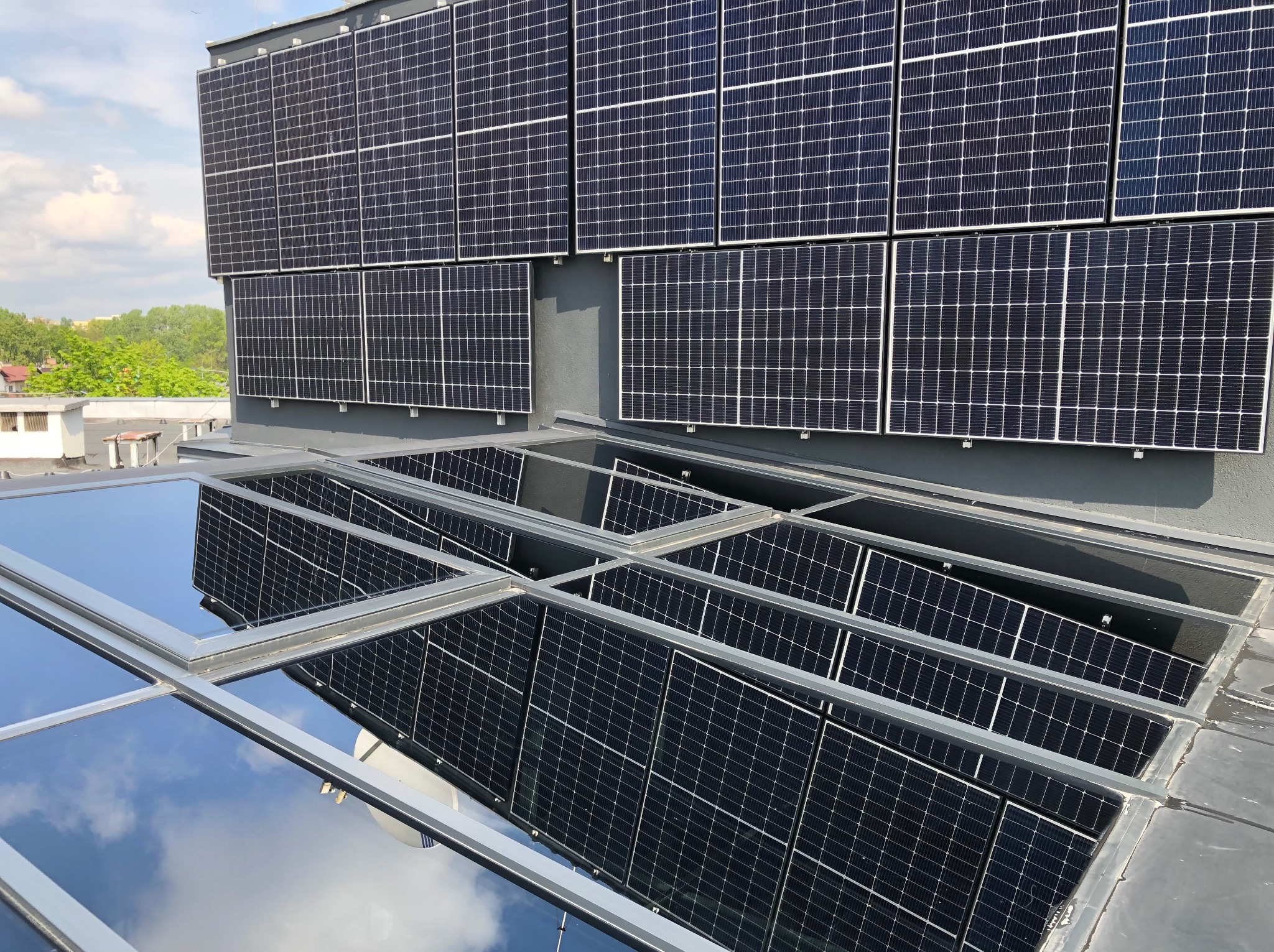 Szary dach budynku z panelami słonecznymi i świetlikami, w których odbija się niebo. Widok z góry, w tle zieleń drzew.