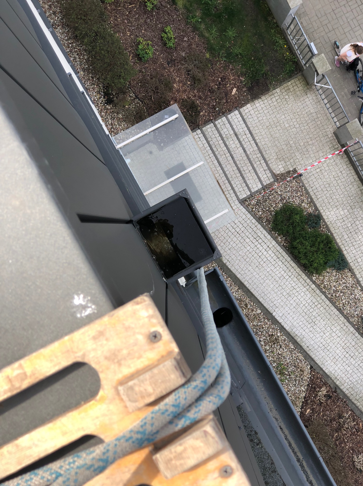 Zablokowana rynna dachowa wypełniona wodą, widok z góry z platformy roboczej. W tle chodnik i zieleń. Prace wysokościowe.