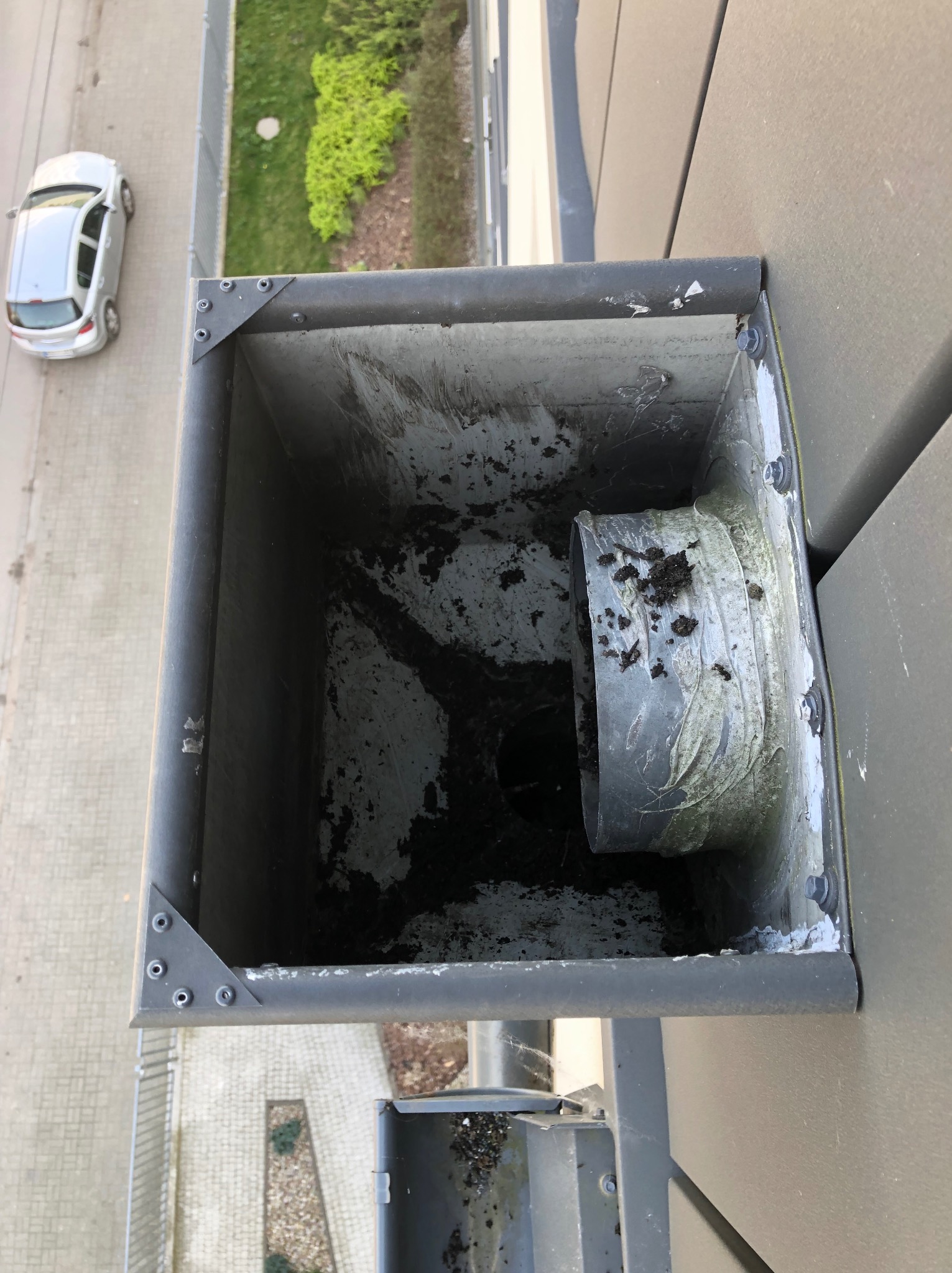 Zanieczyszczona rynna dachowa z widocznymi liśćmi i brudem, sfotografowana z góry. W tle fragment chodnika, trawnika i zaparkowany samochód.
