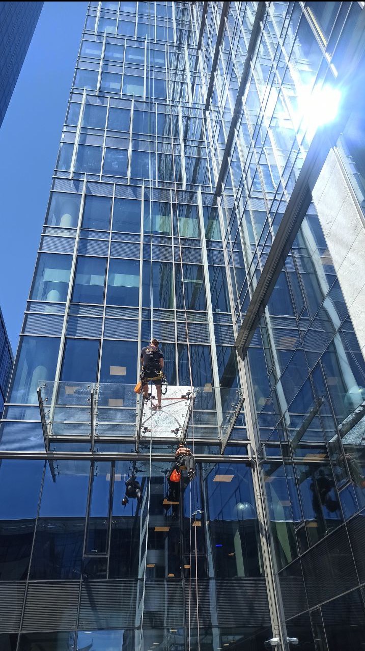 Dwóch alpinistów na platformie myje szklaną fasadę wieżowca w słoneczny dzień. Widok z dołu, podkreślający wysokość i nowoczesną architekturę budynku.