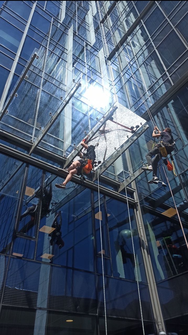 Dwóch alpinistów myje szklaną fasadę biurowca, wisząc na linach. Widok z dołu, słońce odbija się w oknach. Prace wysokościowe.