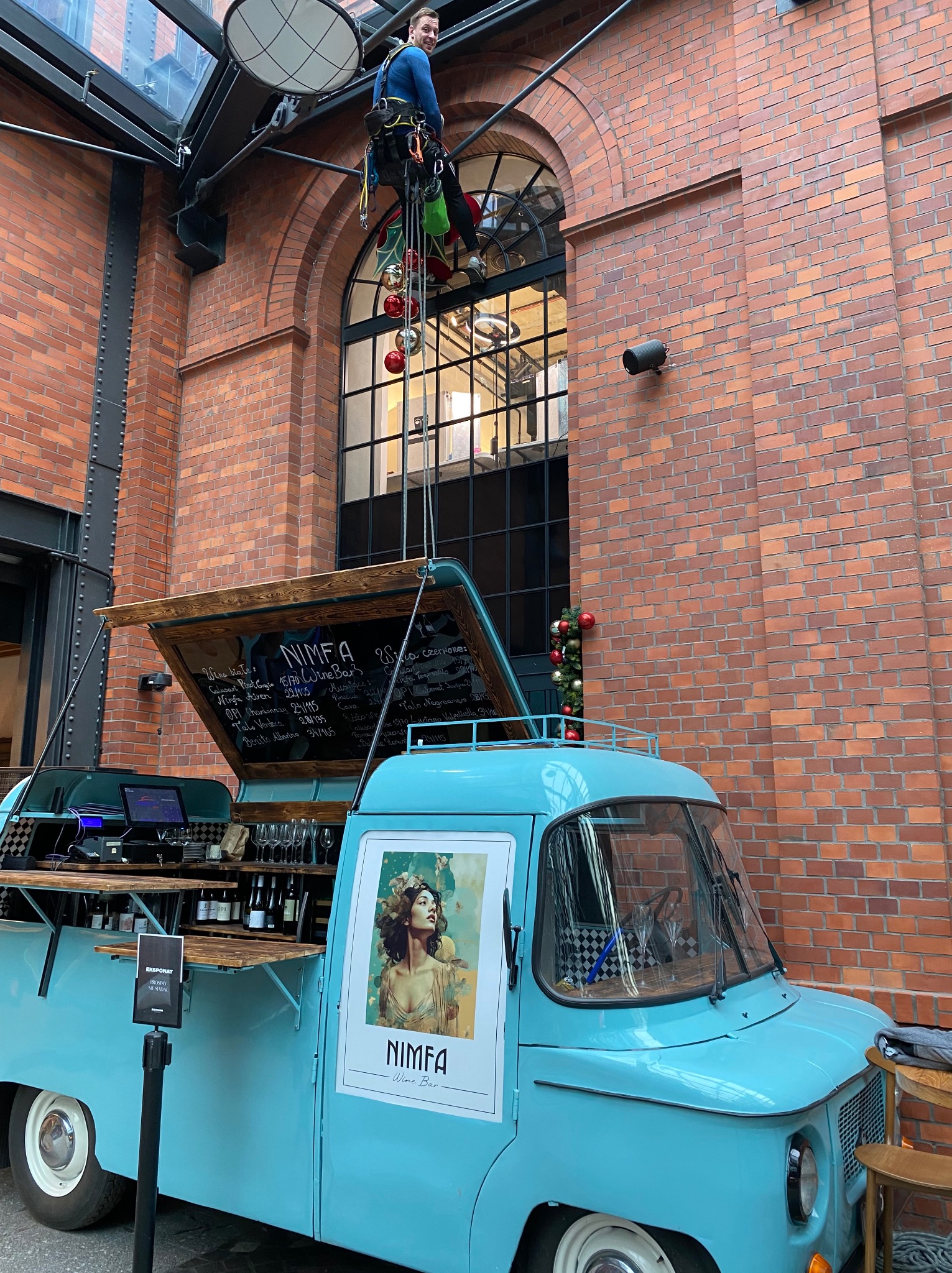 Pracownik na linie, w uprzęży, czyści okna nad mobilnym barem w ceglanym wnętrzu. Dekoracje świąteczne i tablica z menu baru dodają unikalnego charakteru.