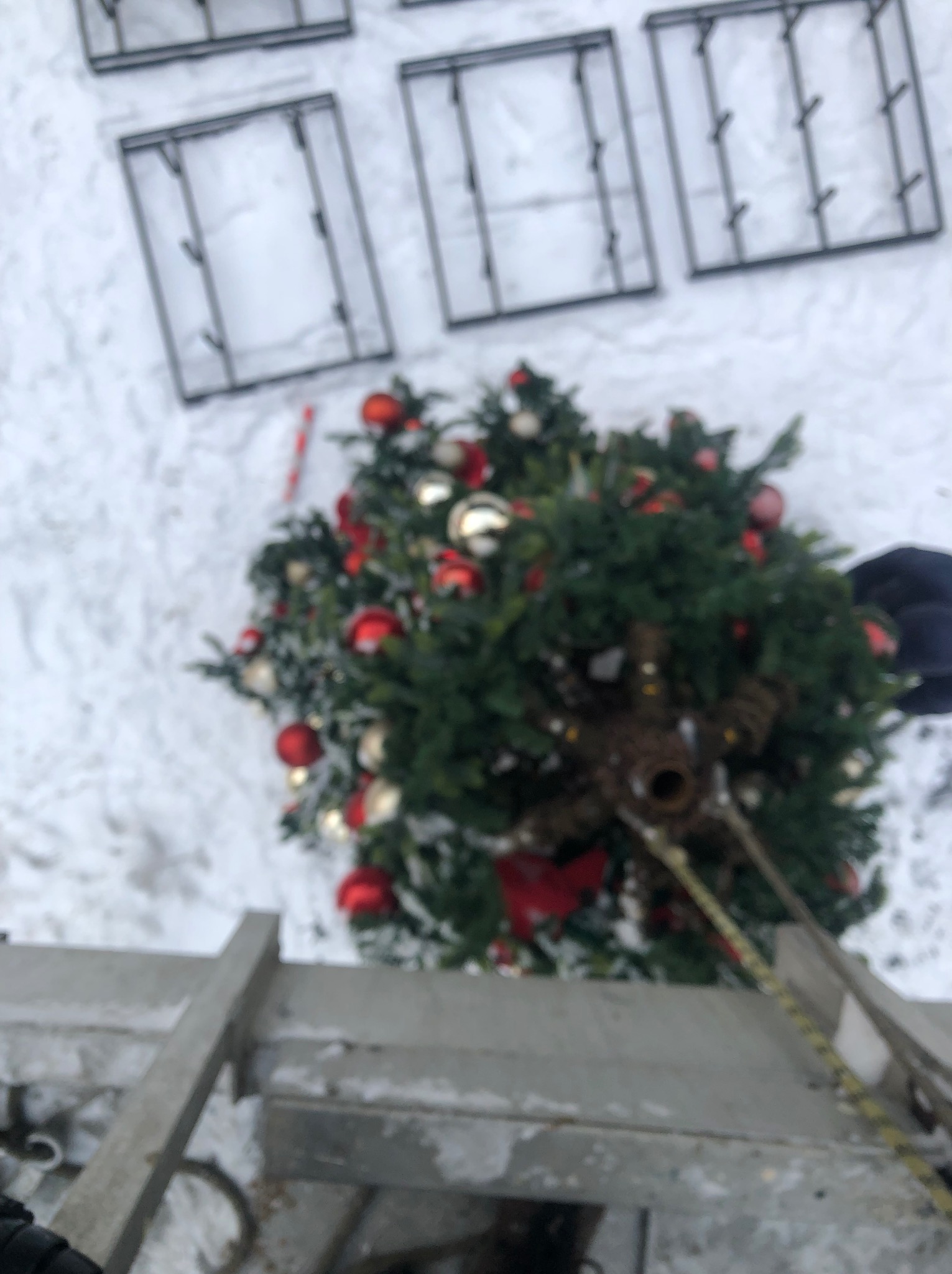 Montaż dekoracji świątecznych na wysokości: choinka z bombkami i drabina na tle zaśnieżonego dachu i okien. Widok z góry, perspektywa pracownika.