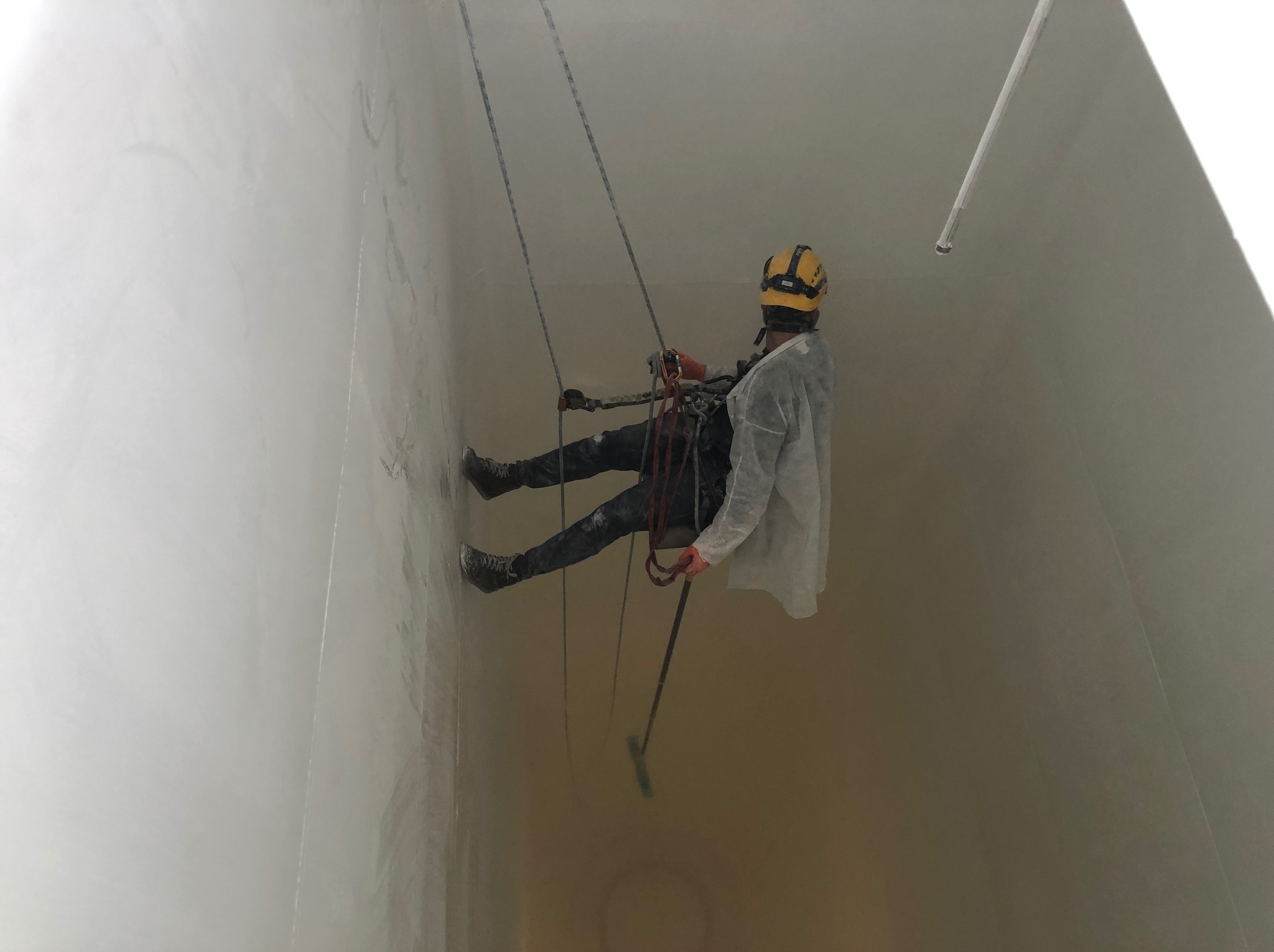 Pracownik w uprzęży, w kasku i kombinezonie, czyści jasną, wysoką ścianę z użyciem narzędzia na długim kiju. Widoczne liny i zabezpieczenia.