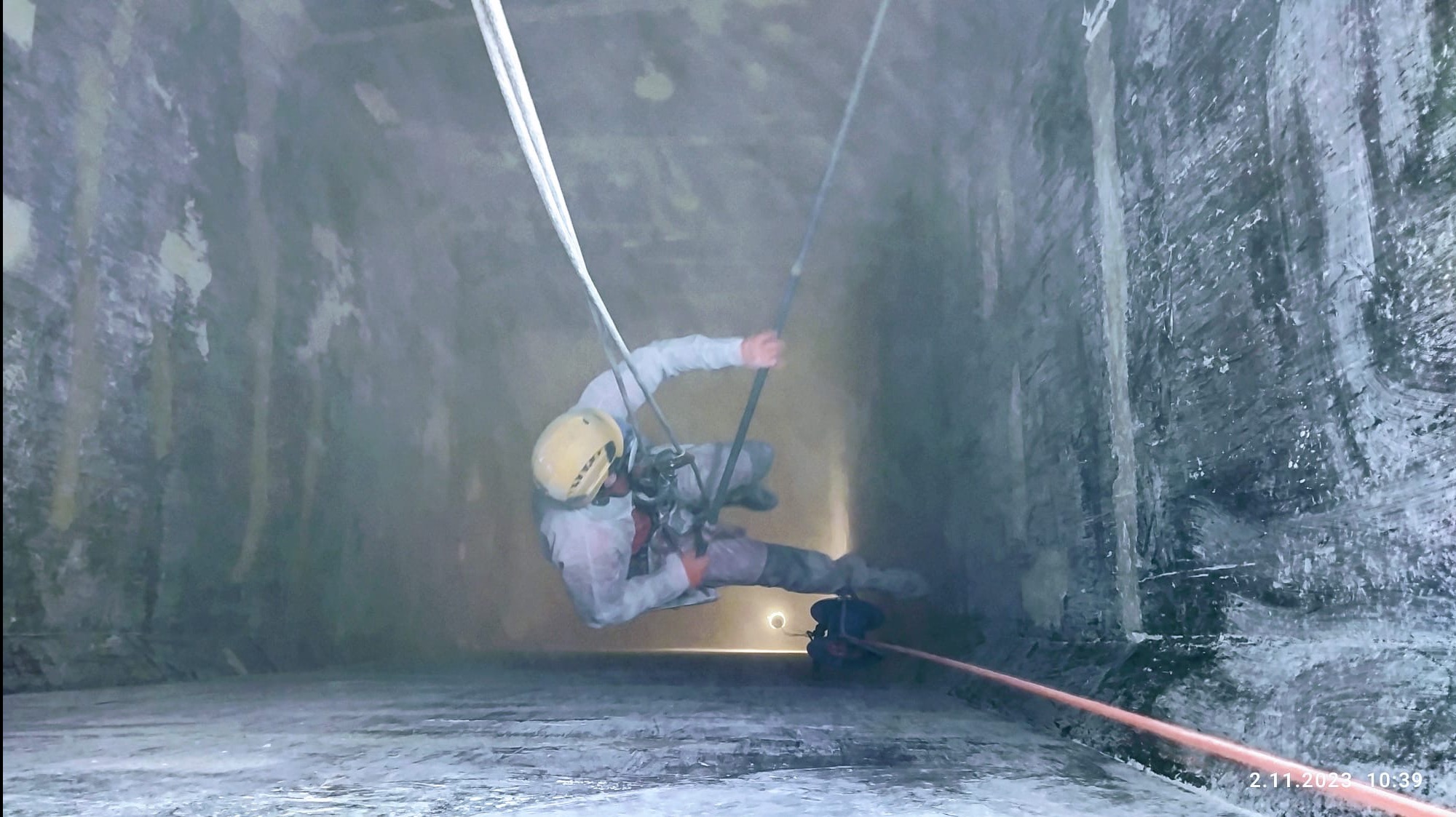 Pracownik w kasku i kombinezonie, zawieszony na linach, wykonuje prace na wysokości wewnątrz betonowej konstrukcji. Widoczne liny, sprzęt asekuracyjny i oświetlenie.