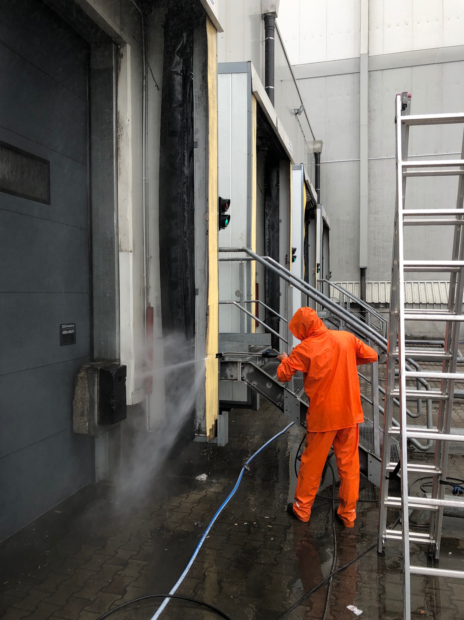 Pracownik w pomarańczowym kombinezonie czyści elewację hali przemysłowej myjką ciśnieniową. Widoczna drabina i fragmenty elewacji z rampami załadunkowymi.