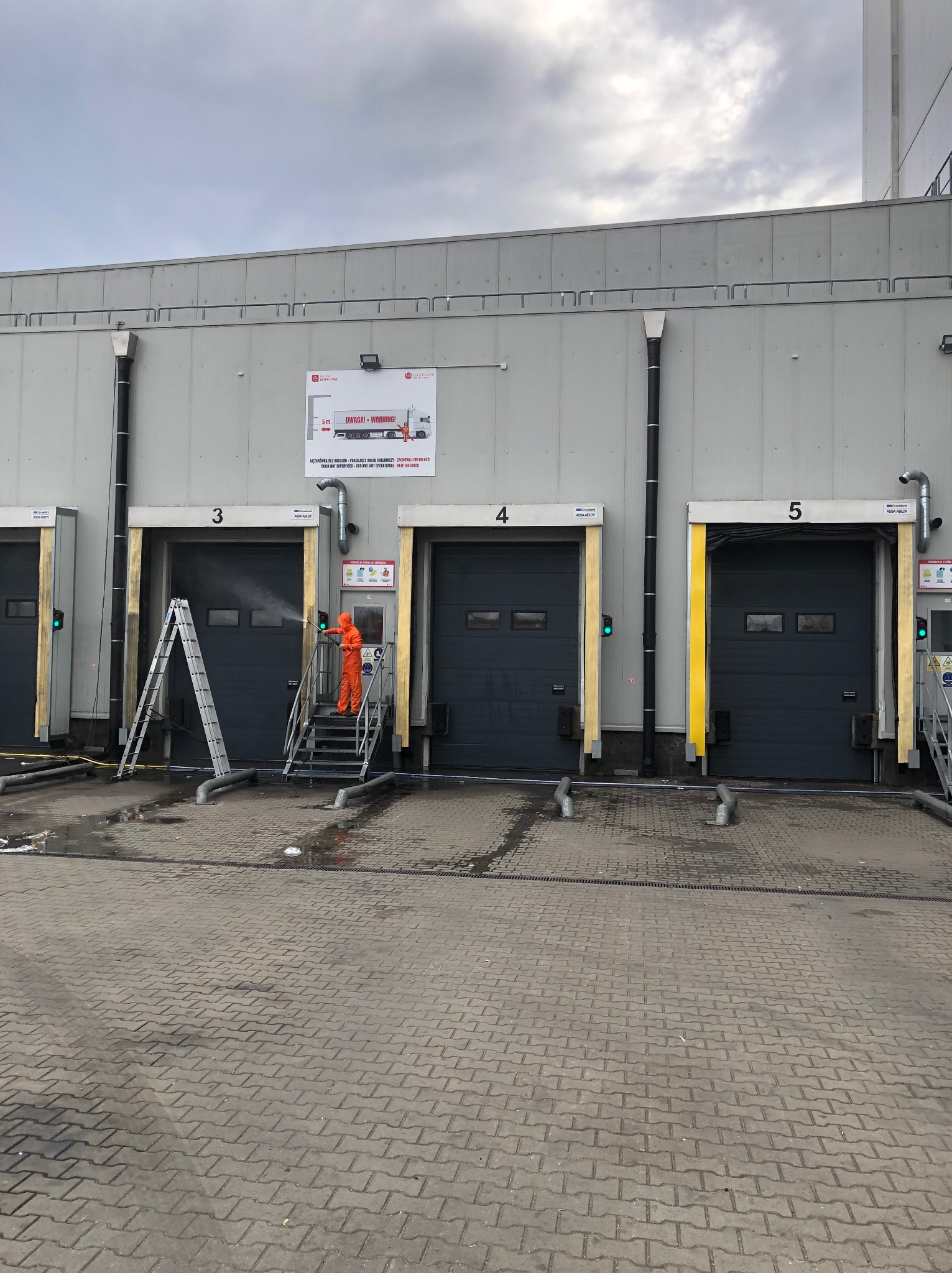 Mycie bram przemysłowych z użyciem myjki ciśnieniowej przez pracownika w kombinezonie ochronnym. Widoczne trzy bramy, drabina i woda pod ciśnieniem.