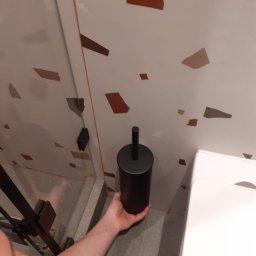 G&O - Nowoczesna łazienka z mozaikową ścianą i czarnym pojemnikiem na szczotkę WC trzymanym w dłoni, obok widoczna biała toaleta i szklana kabina prysznicowa.