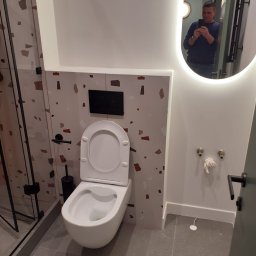 G&O - Nowoczesna toaleta wisząca w łazience z mozaiką, widoczne przyłącza wody i odbicie osoby w podświetlanym lustrze. Minimalistyczny design.