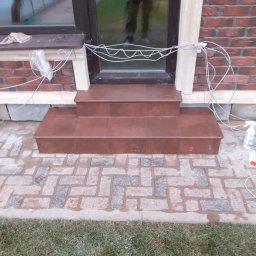 G&O - Wykończone płytkami schody wejściowe do domu, z widoczną kostką brukową i fragmentem trawnika. Ujęcie z poziomu gruntu, z przewodami i środkami czystości w tle.