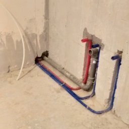 G&O - Surowy kąt łazienki z instalacją wodną: rury miedziane i plastikowe (czerwone i niebieskie) wystają ze ściany, przygotowane do podłączenia armatury. Widoczne ślady po kuciu.