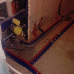 G&O - Instalacja wodno-kanalizacyjna w trakcie montażu: rury PEX w izolacji czerwono-niebieskiej, szara rura kanalizacyjna, elementy mocujące do ściany. Surowy stan.