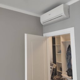G&O - Klimatyzator Panasonic zamontowany nad wejściem do garderoby z półkami. Ściana pomalowana na szaro, białe listwy przysufitowe i ościeżnica. Widoczne ubrania w szafie.