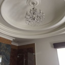 Biały sufit z dekoracyjnym gzymsem i kryształowym żyrandolem w łazience z marmurowymi ścianami i kabiną prysznicową. Eleganckie wykończenie wnętrza.