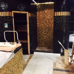 Sauna w trakcie wykańczania: czarne mozaiki z złotymi akcentami, drewniane elementy i narzędzia pracy. Widoczny etap prac wykończeniowych.