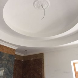 Biały, okrągły sufit podwieszany z dekoracyjnym elementem centralnym i wystającym przewodem. Fragment ściany wykończony mozaiką i płytami imitującymi marmur.