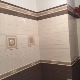 G&O - Wykończona łazienka z brązowo-białymi płytkami, dekoracyjnymi bordiurami i ozdobnymi ramkami na ścianie. Widoczny fragment sufitu z listwą. Na dole leży pędzel.