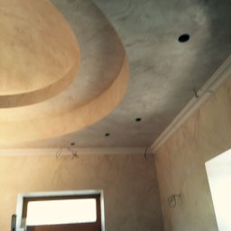 G&O - Kremowo-szary sufit z dekoracyjnym gzymsem i okrągłym wgłębieniem, widok z dołu. W tle fragment okna i ściany z fakturą. Widoczne przewody elektryczne.