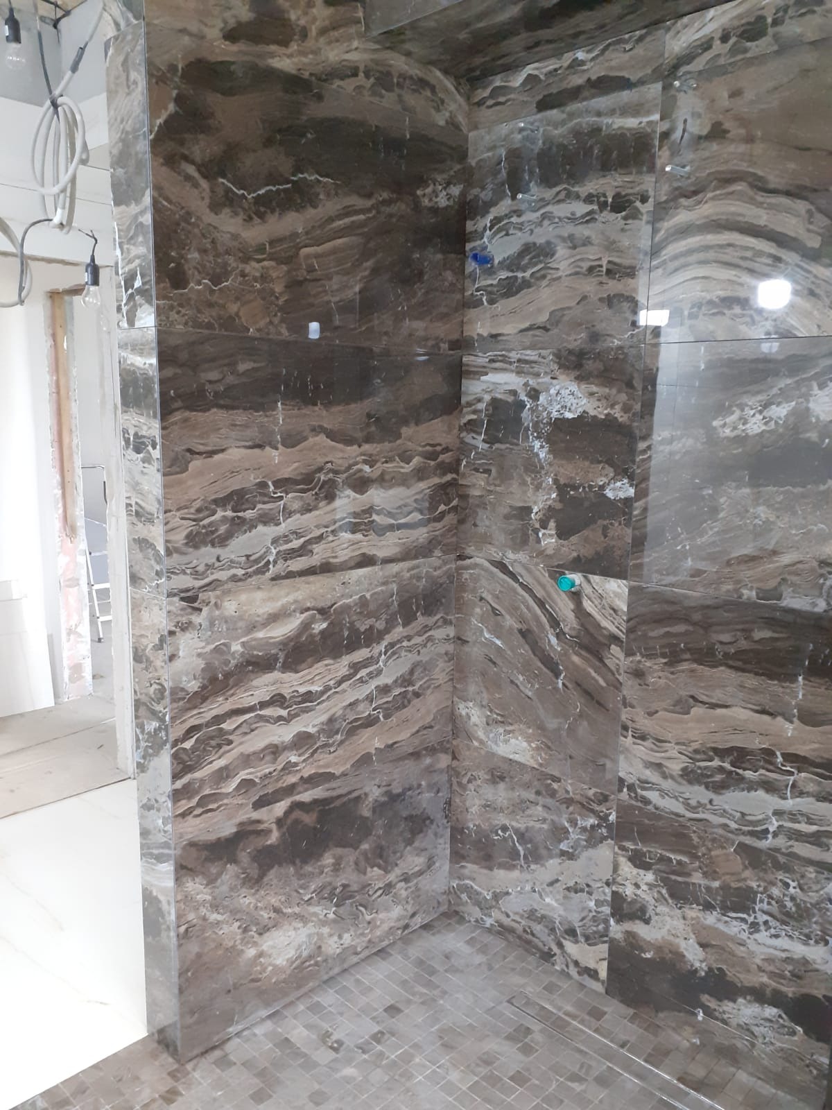 Wykończona kabina prysznicowa z brązowymi płytkami imitującymi marmur, z widocznym odpływem liniowym i fragmentem instalacji elektrycznej w tle.