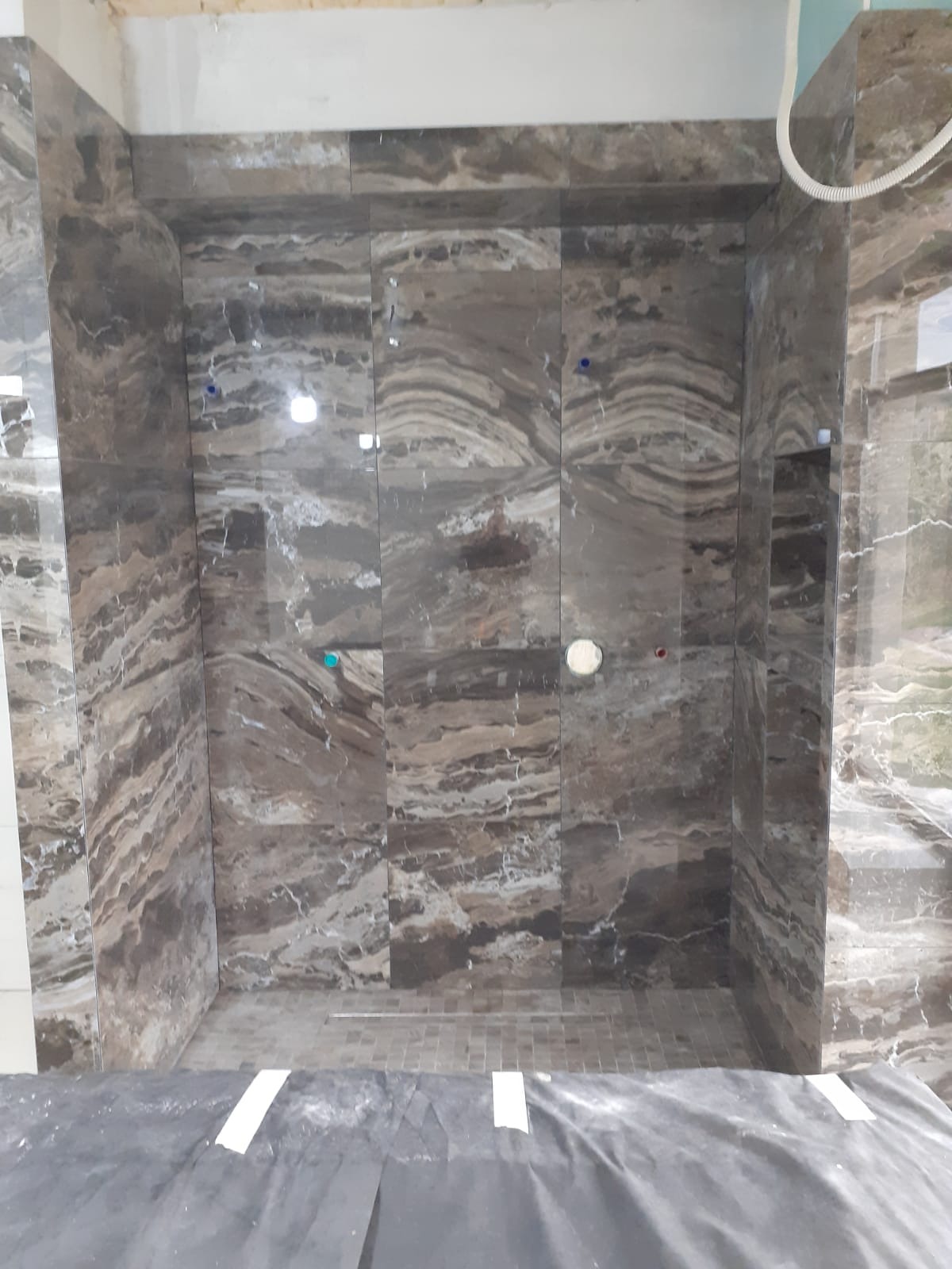 Kabina prysznicowa w trakcie wykańczania, z brązowymi płytkami imitującymi marmur na ścianach i mozaiką na podłodze. Widoczne elementy instalacji i zabezpieczenia podłogi.