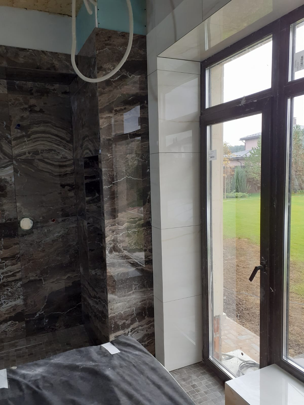 Częściowo wykończona łazienka z marmurowymi i białymi płytkami, widok na zewnątrz przez okno. Podłoga zabezpieczona folią ochronną.