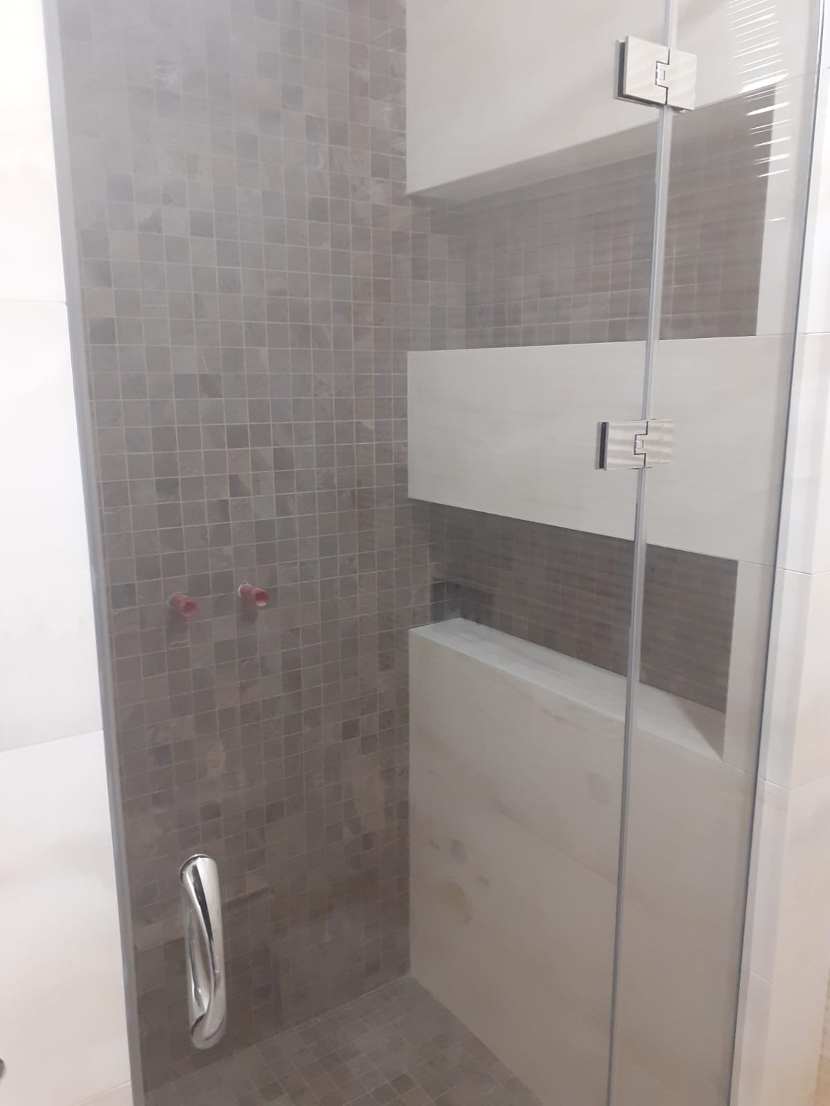 Szklana kabina prysznicowa z mozaiką w odcieniach szarości i białymi panelami. Widoczne chromowane okucia i uchwyt. Minimalistyczny design.