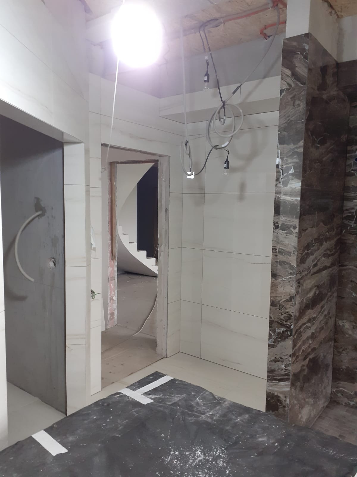 Remont łazienki w trakcie: ułożone płytki imitujące marmur na ścianie, widoczne kable oświetleniowe, zabezpieczona podłoga. W tle widoczne schody.