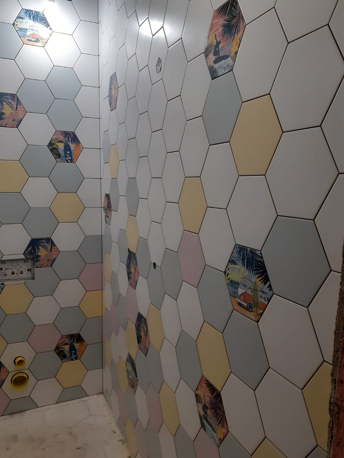 Ściana łazienki wyłożona heksagonalnymi płytkami w pastelowych kolorach z motywami plażowymi, widoczne fragmenty instalacji wodnej. Nietypowy wzór płytek.