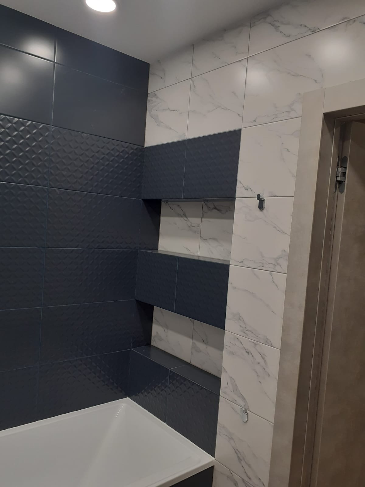 Nowoczesna łazienka z ciemnymi, teksturowanymi płytkami i białymi płytkami imitującymi marmur. W ścianie wnęki wykończone płytkami. Fragment wanny.