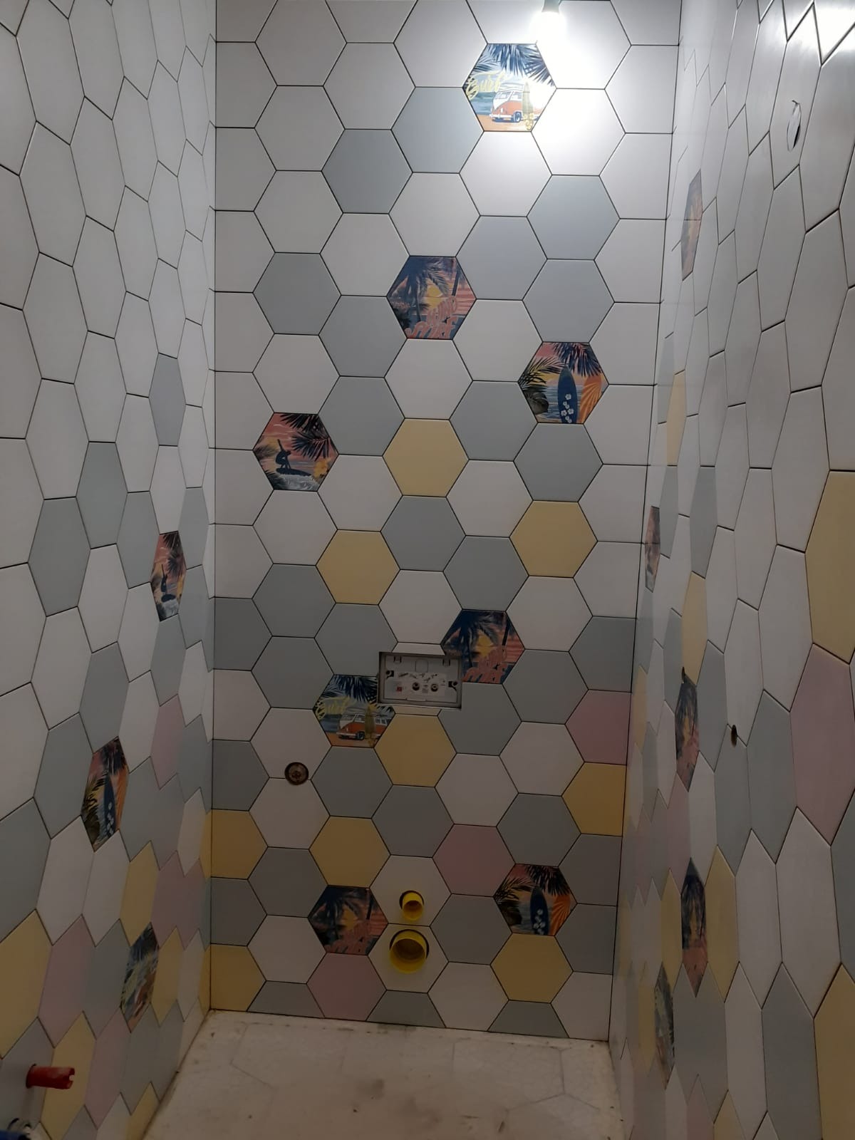 Łazienka z heksagonalnymi płytkami w odcieniach szarości, żółci i bieli, z motywami plażowymi. Widoczne elementy instalacji wodnej i stelaż podtynkowy. Nietypowy wzór płytek.