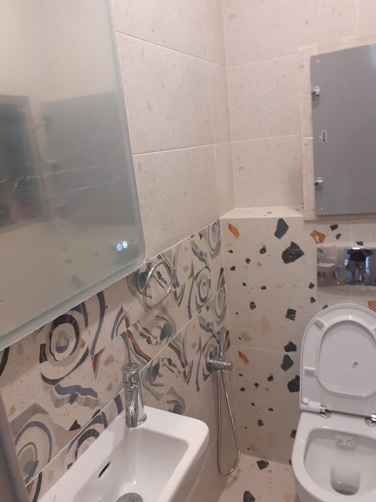 Nowoczesna łazienka z umywalką, toaletą i podświetlanym lustrem. Uwagę przykuwają oryginalne płytki w stylu lastryko i dekoracyjne wzory na dolnej części ścian.