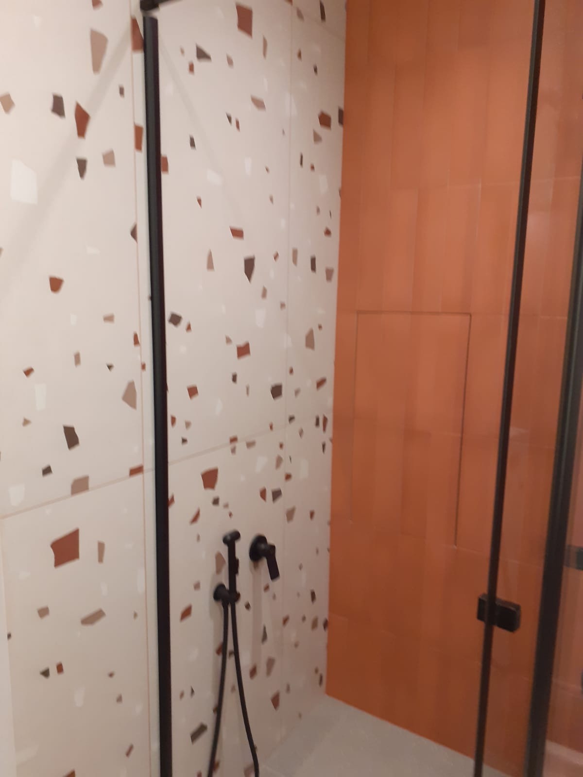 Nowoczesna kabina prysznicowa z czarną armaturą i płytkami w stylu lastryko na ścianach, kontrastująca z pomarańczową ścianą. Detal wykończenia łazienki.