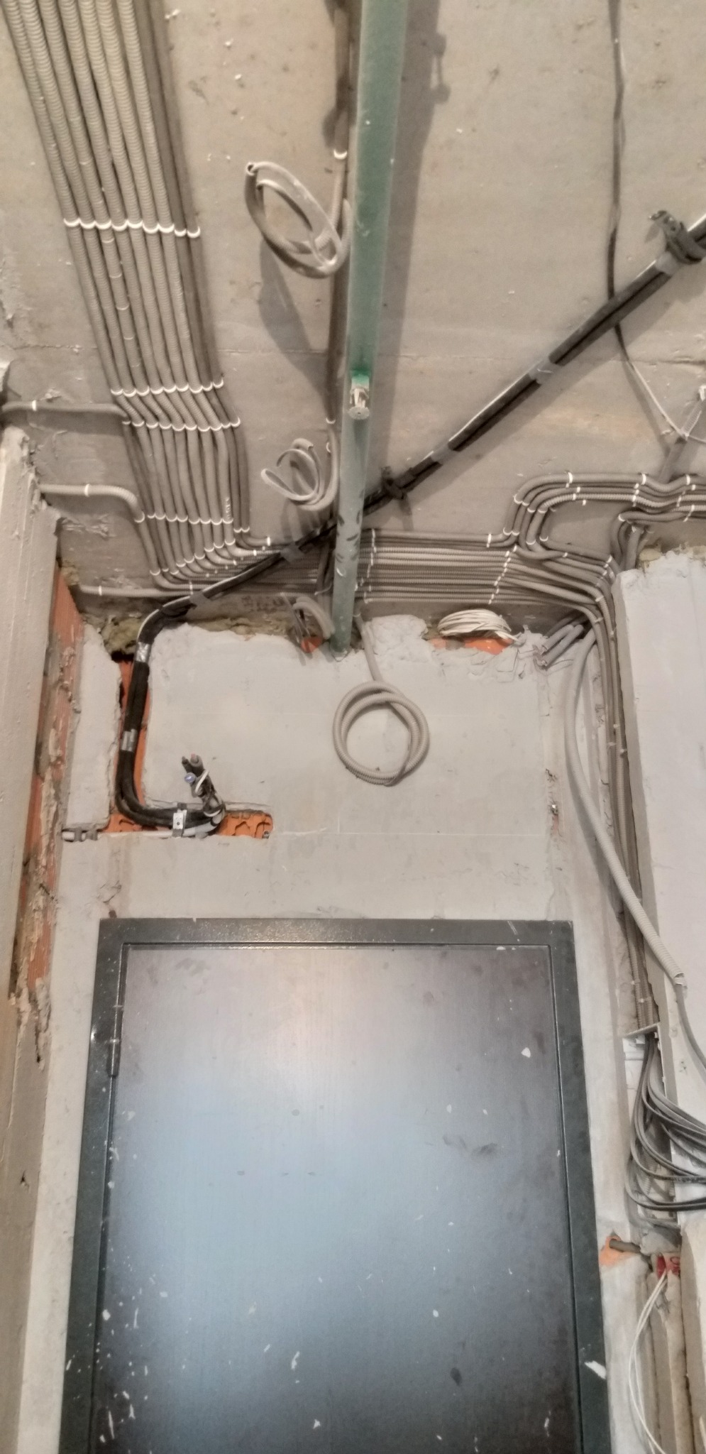 Instalacja elektryczna w peszlach na suficie i ścianie, widoczne kable i rury, surowy stan wykończenia, otwór w ścianie, metalowe drzwi.
