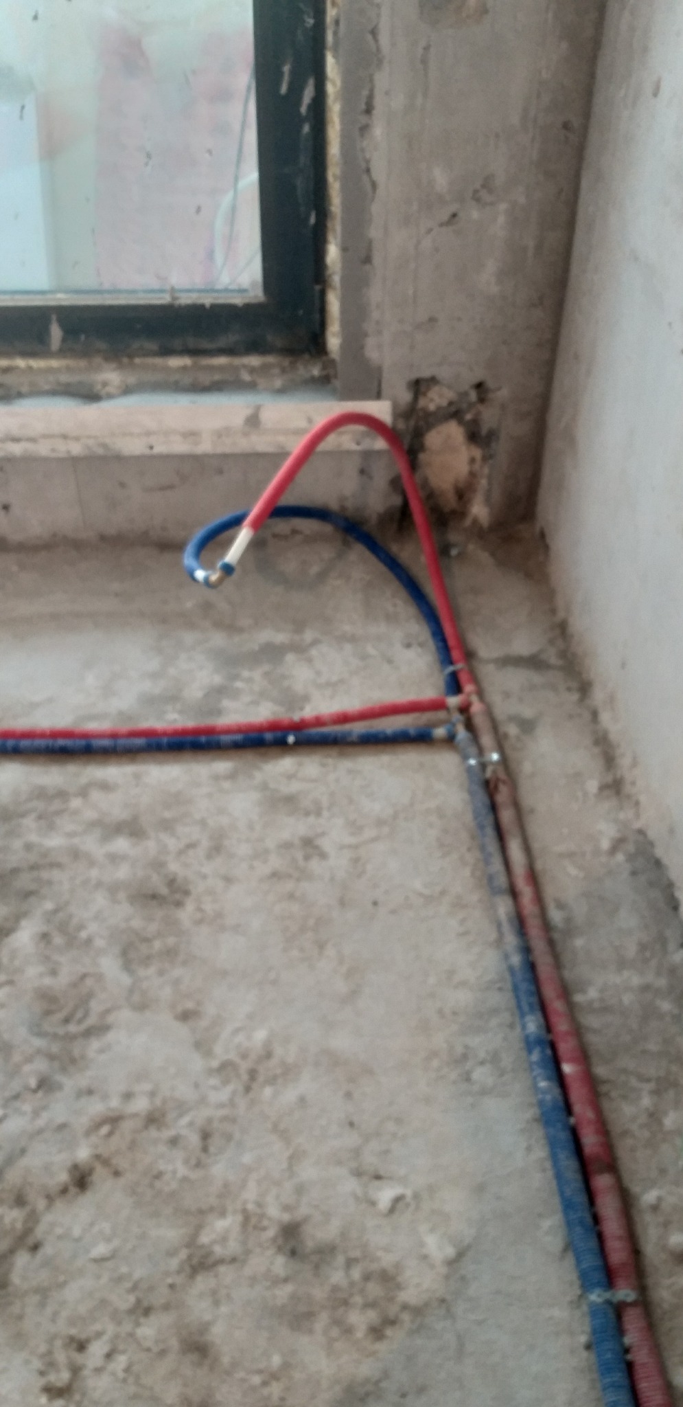 Instalacja ogrzewania podłogowego w trakcie prac: czerwone i niebieskie rurki ułożone na surowym podłożu, widoczne przyłącza i fragment okna.