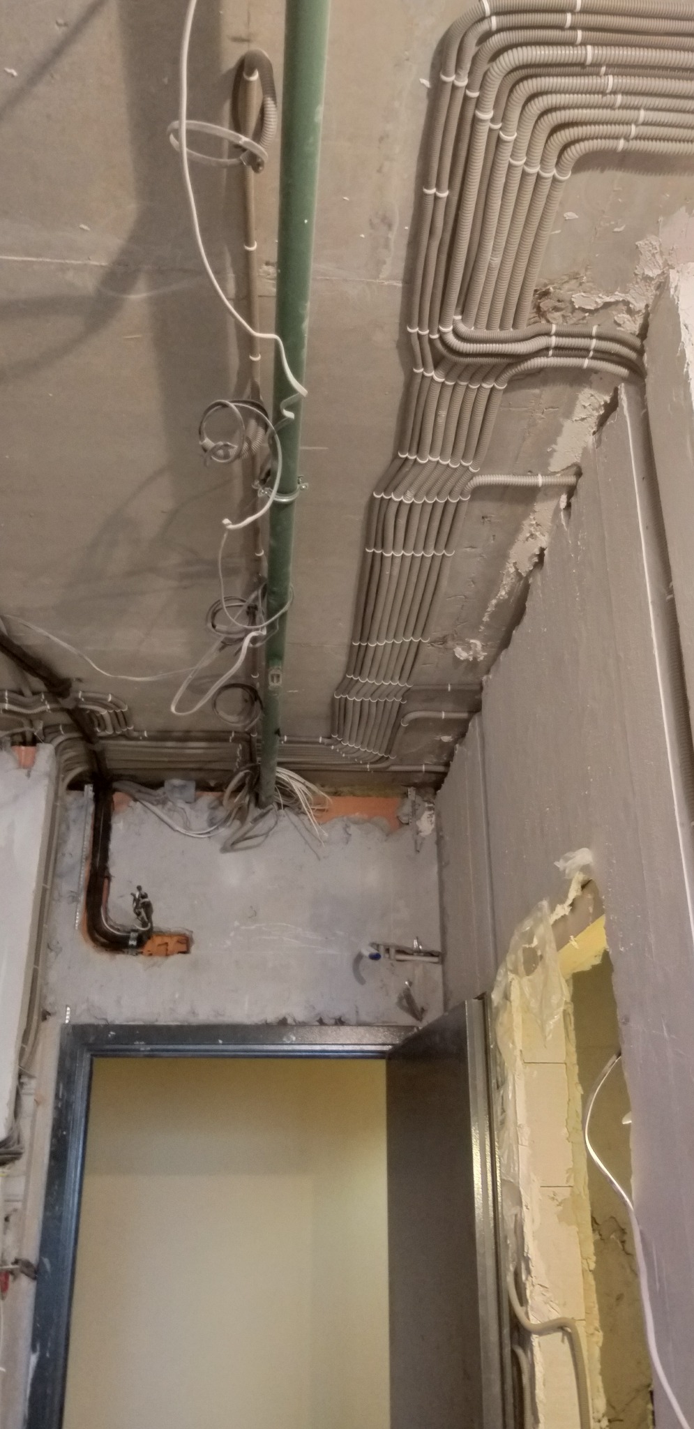 Instalacja elektryczna w peszlach ułożona pod sufitem, widoczne przewody i rury. Stan surowy, przygotowanie do dalszych prac. Widok na futrynę drzwi.