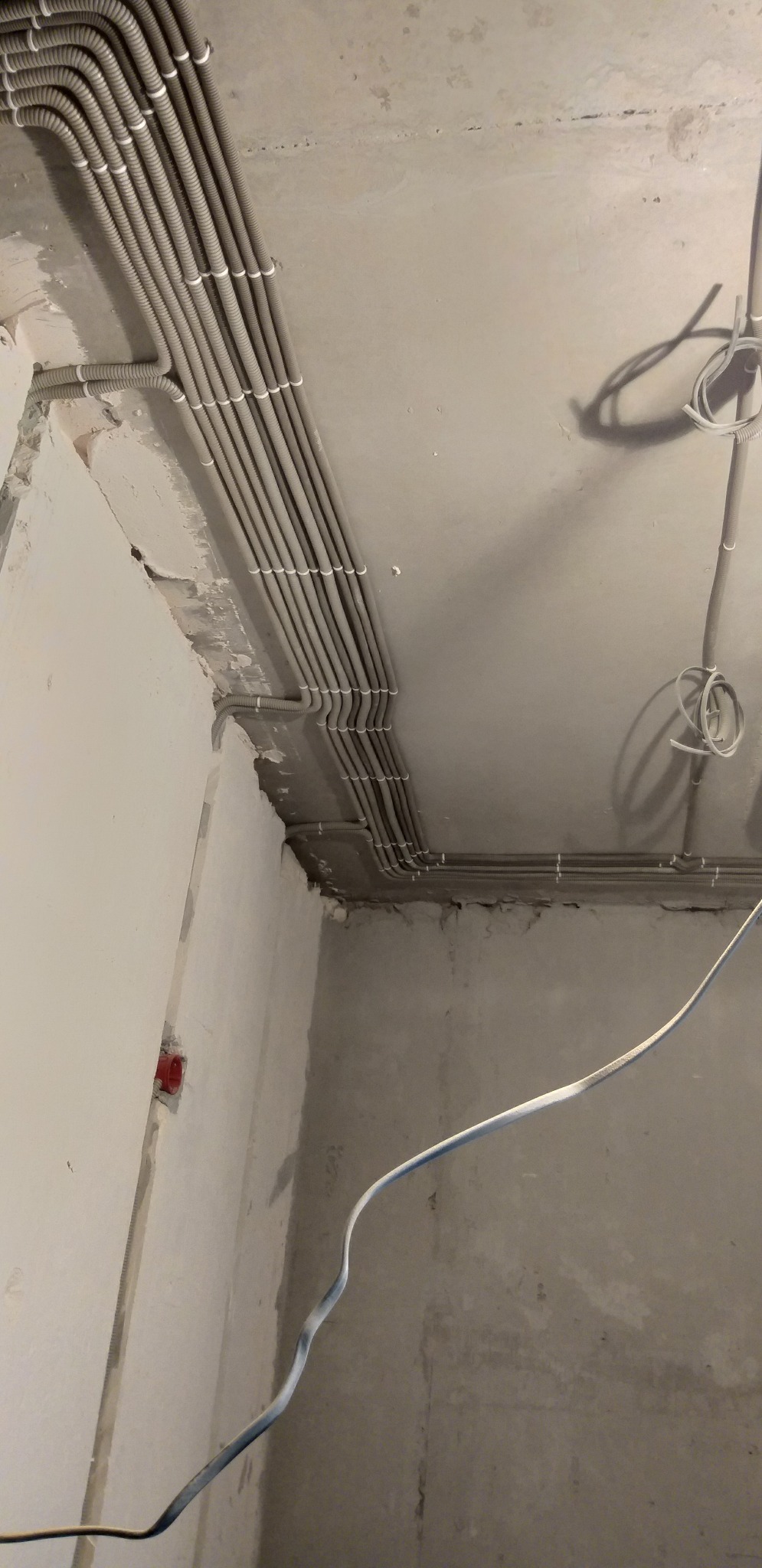 Szare rury instalacji elektrycznej ułożone równolegle pod sufitem w surowym wnętrzu. Widoczne kable i puszka elektryczna w ścianie. Ujęcie z dołu, surowy stan.