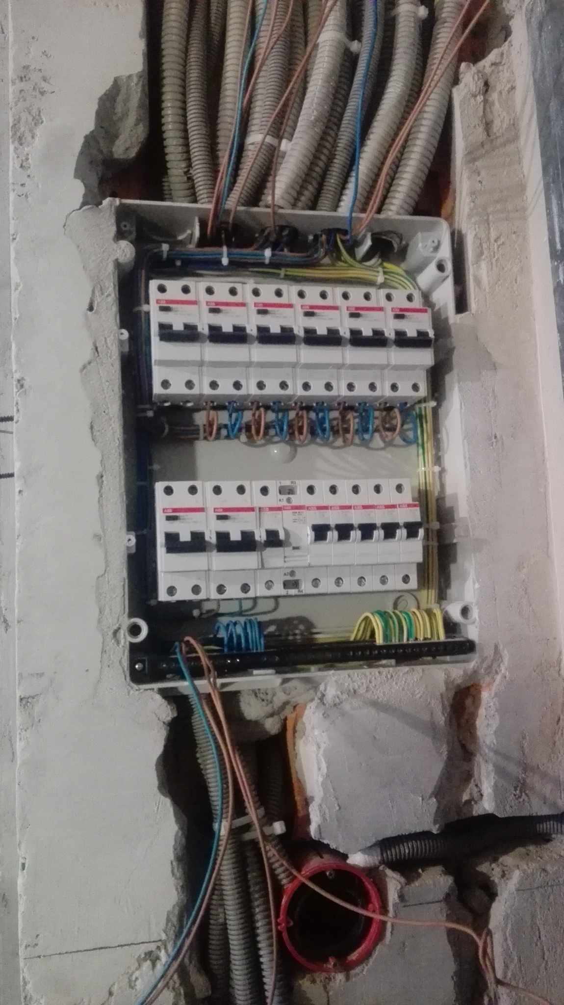 Rozdzielnia elektryczna z zabezpieczeniami w surowym stanie, widoczne kable w peszlach i przewody. Montaż instalacji elektrycznej w trakcie wykończenia wnętrza.