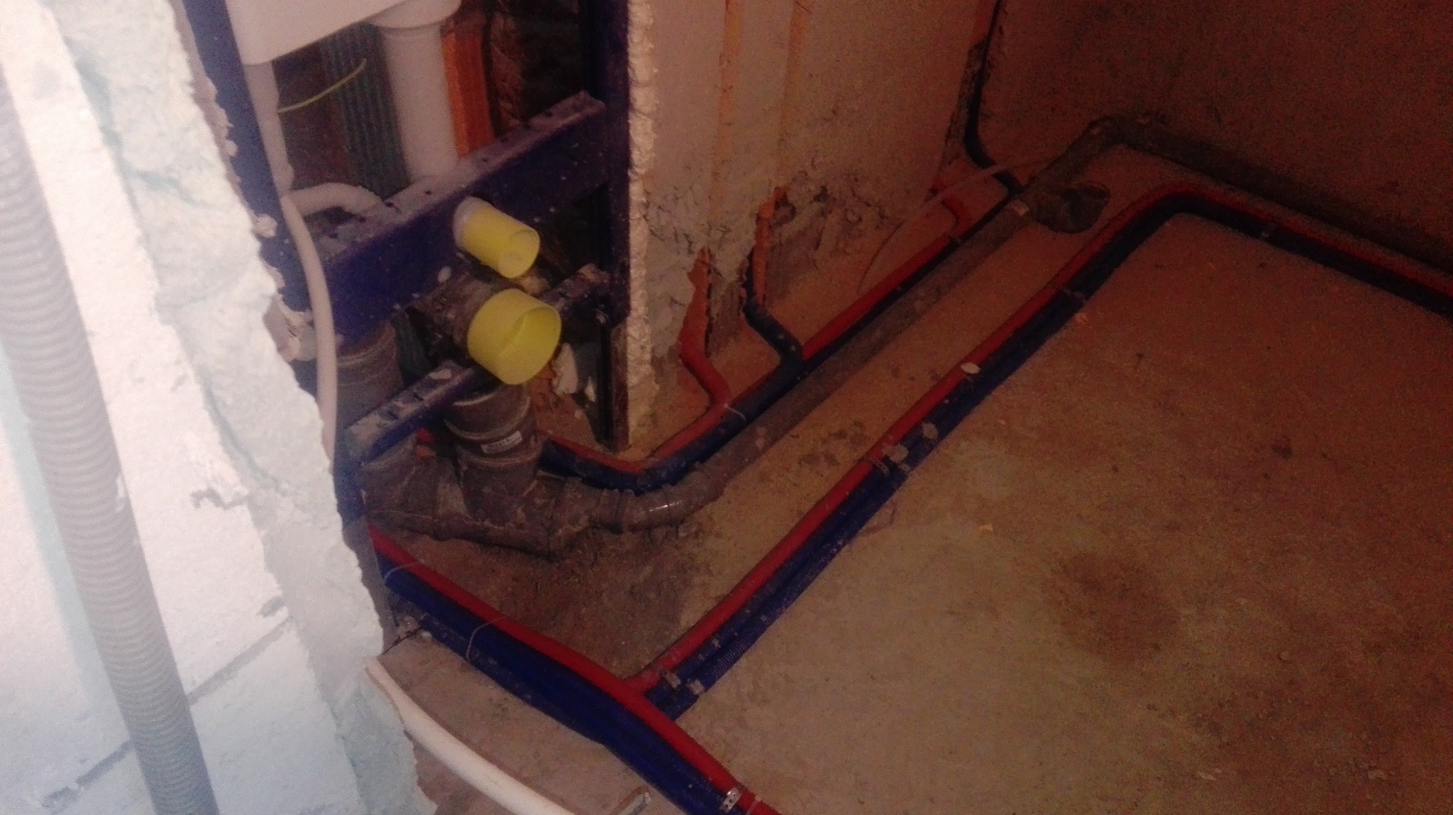 Instalacja wodno-kanalizacyjna w trakcie montażu: rury PEX w izolacji czerwono-niebieskiej, szara rura kanalizacyjna, elementy mocujące do ściany. Surowy stan.